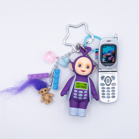 A 90s-themed keychain featuring a purple and blue Tinky Winky figure with a Bandai mini flip phone and charms including a cassette, blue and purple lego pieces, a pink dice, and a troll with purple hair
