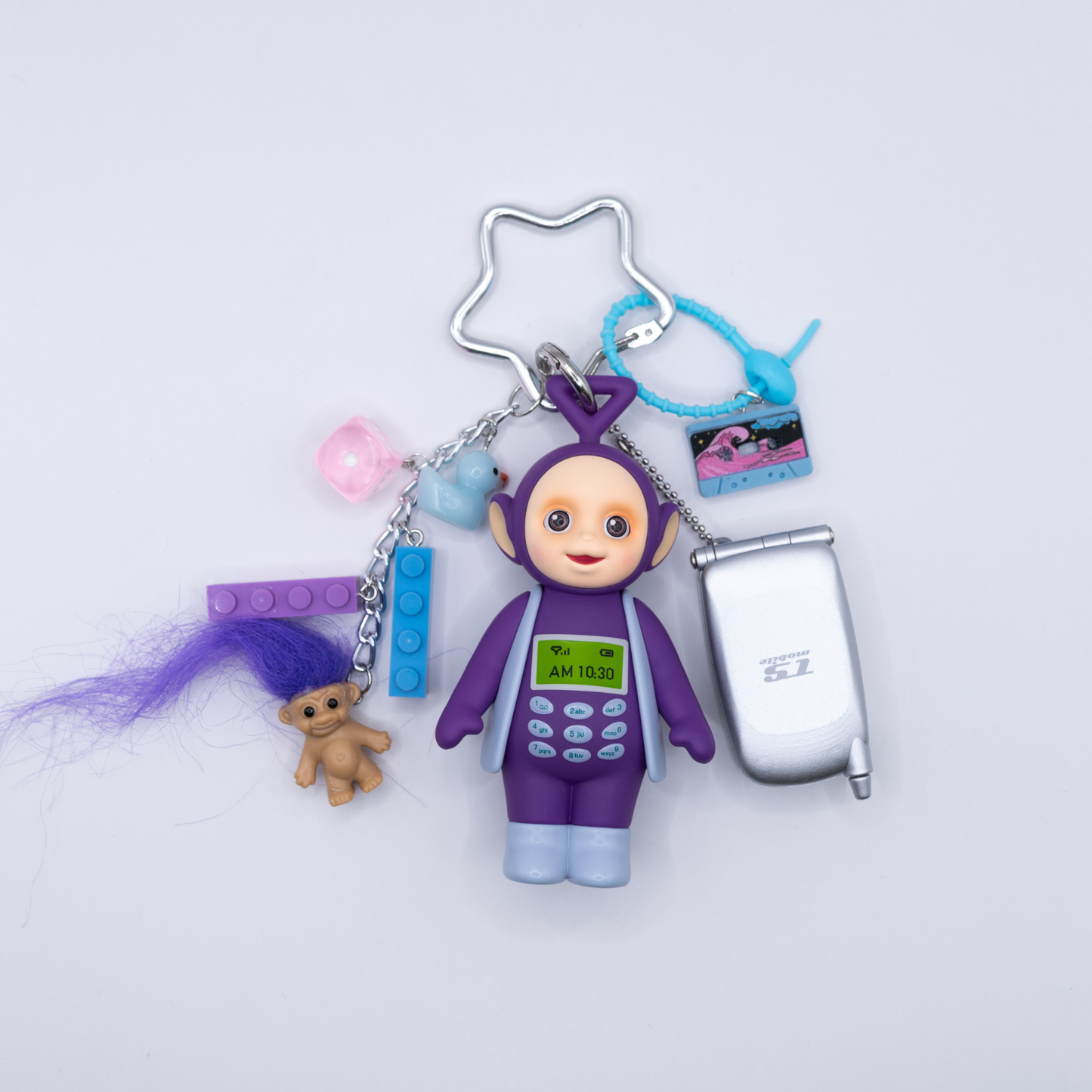 A 90s-themed keychain featuring a purple and blue Tinky Winky figure with a Bandai mini flip phone and charms including a cassette, blue and purple lego pieces, a pink dice, and a troll with purple hair