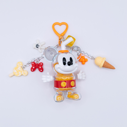 A Mickey Mouse-themed acrylic charm keychain featuring a clear Mickey figure in a yellow and red outfit, surrounded by small dangling charms including a lollipop, bow tie, ice cream cone, and heart-shaped clip, photographed on a clean white background.