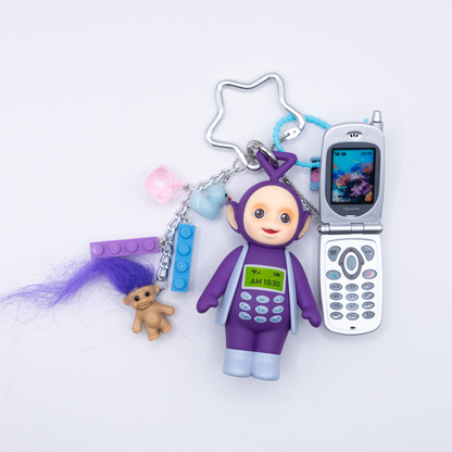 A 90s-themed keychain featuring a purple and blue Tinky Winky figure with a Bandai mini flip phone and charms including a cassette, blue and purple lego pieces, a pink dice, and a troll with purple hair