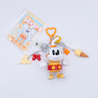 A Mickey Mouse-themed keychain charm set featuring a clear plastic Mickey figure in a red and yellow clown outfit, with dangling accessories including an orange heart-shaped ring, yellow bow, red bow, ice cream cone, and a small yellow charm. A printed Mickey card is visible behind the keychain on a light gray background.