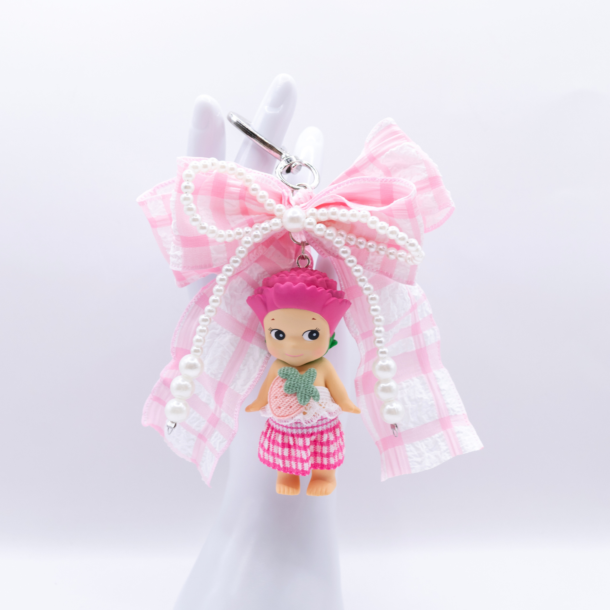 Silver heart keychain featuring Sonny Angel Carnation figure with a pink gingham bow and faux pearls on a white background