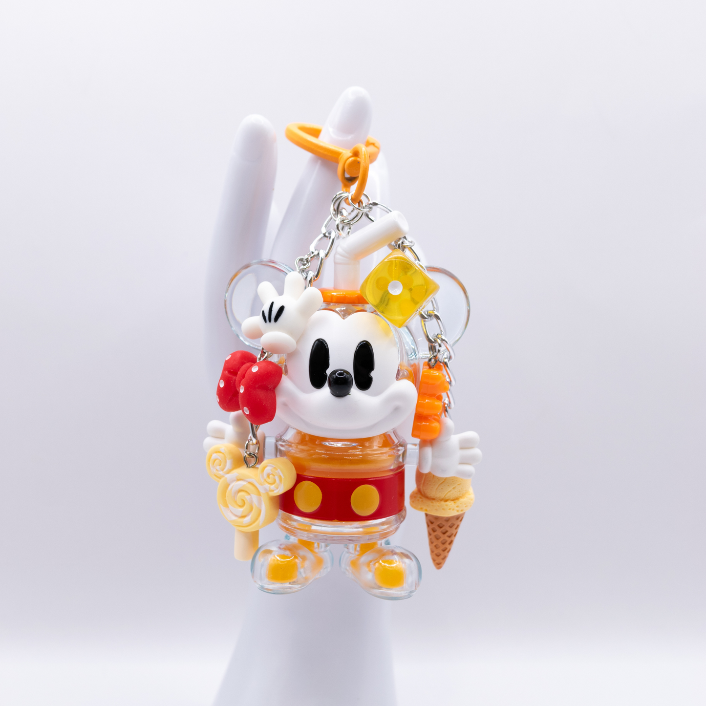 A dangling Mickey Mouse-themed acrylic charm keychain featuring a clear Mickey figure in a yellow and red outfit, surrounded by small dangling charms including a lollipop, bow tie, ice cream cone, and heart-shaped clip, photographed on a clean white background.