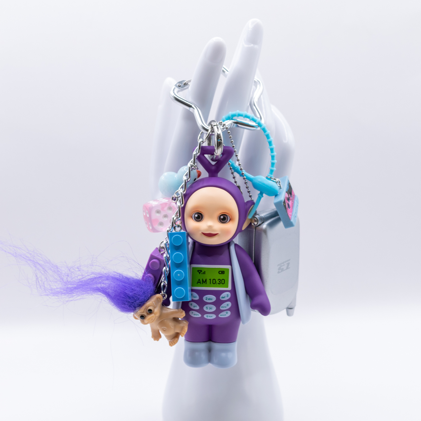 A 90s-themed keychain featuring a purple and blue Tinky Winky figure with a Bandai mini flip phone and charms including a cassette, blue and purple lego pieces, a pink dice, and a troll with purple hair