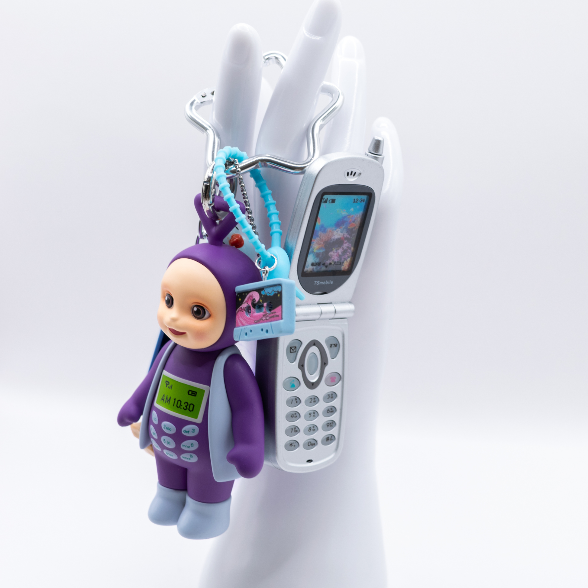 A 90s-themed keychain featuring a purple and blue Tinky Winky figure with a Bandai mini flip phone and charms including a cassette, blue and purple lego pieces, a pink dice, and a troll with purple hair
